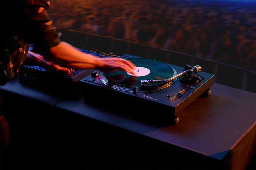 Tocadiscos Technics SL-1210MKX negro con brazo plateado sobre mesa de cabina y mano de un DJ sobre el disco